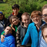 Partecipanti e operatori del Centro Socio Riabilitativo Diurno di Podenzano sorridono insieme durante il soggiorno a Carisolo, in un paesaggio naturale tra boschi, montagne e il torrente